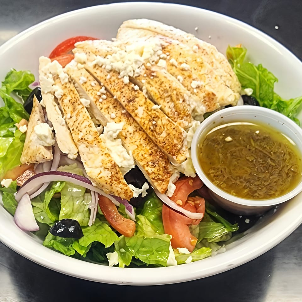 Grilled chicken salad with feta, olives, tomatoes, red onion, and dressing in a white bowl.