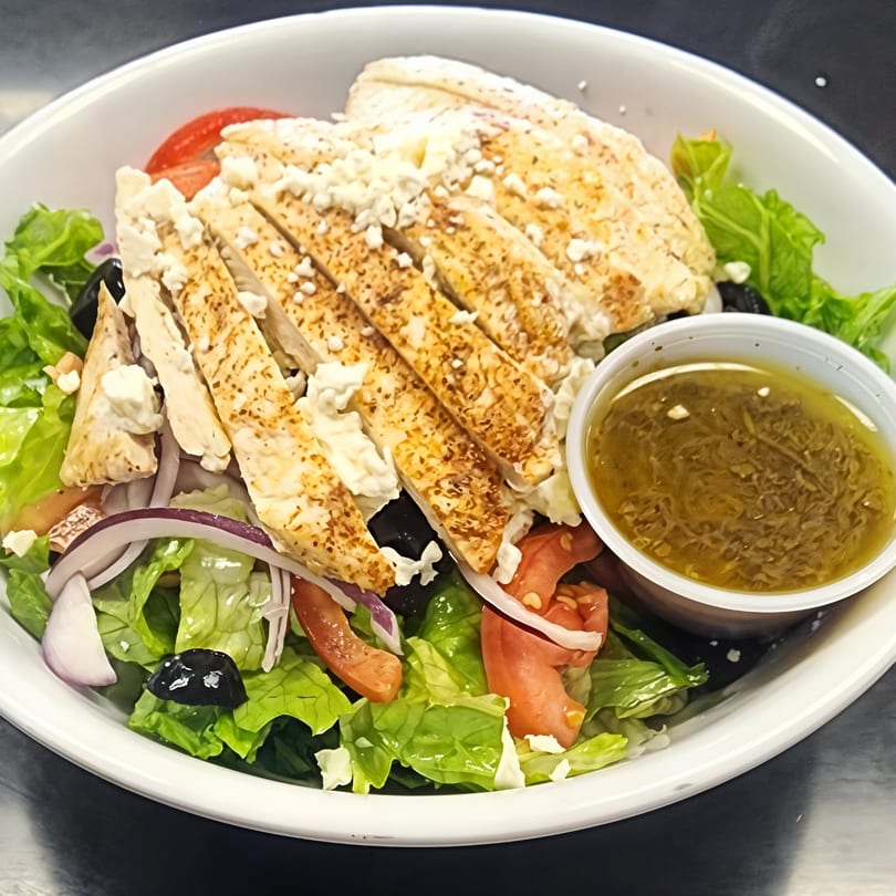 Grilled chicken salad with feta, olives, tomatoes, red onion, and dressing in a white bowl.